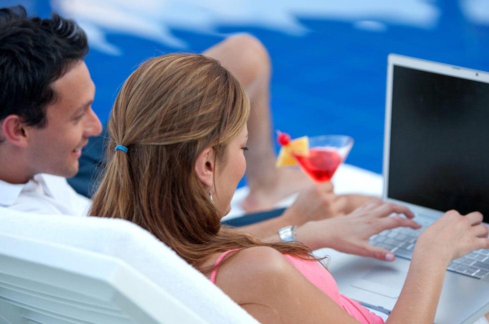 A man and woman sit on lounge chairs by the pool, using a laptop and enjoying cocktails.