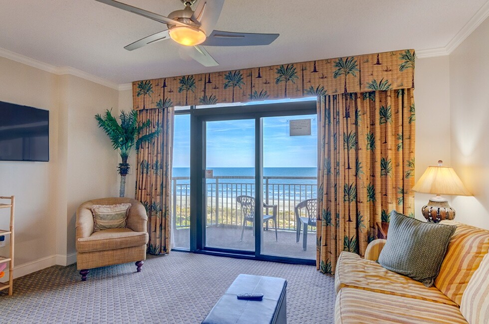 A wide view of a hotel room with a sofa, chair, coffee table, TV, and balcony with an ocean view.