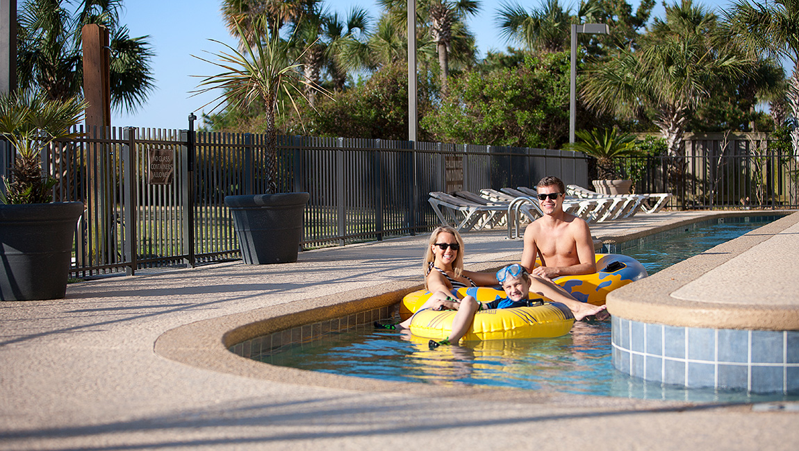 A family of three enjoys a sunny day in a swimming pool with inflatable toys.