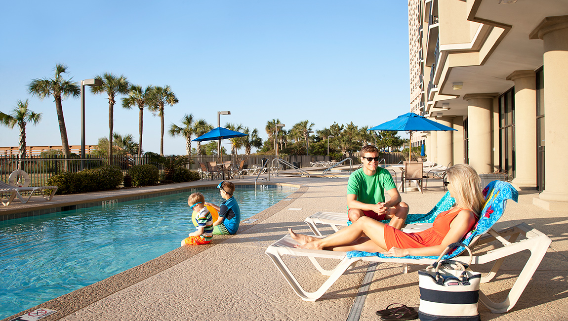 A family enjoys the outdoor pool area of a hotel with a beige building in the background.
